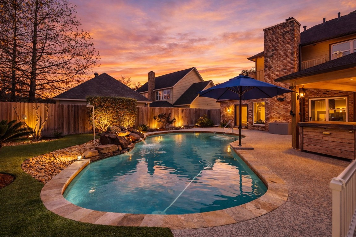 6618 Wimbledon Trail Road Spring, TX 77379 - Photo 37 of 43 This stunning backyard features a beautifully lit swimming pool with a natural rock waterfall, surrounded by lush landscaping. A cozy patio area and outdoor lighting creates a perfect setting for evening relaxation. The backdrop of a warm sunset enhances the inviting ambiance.