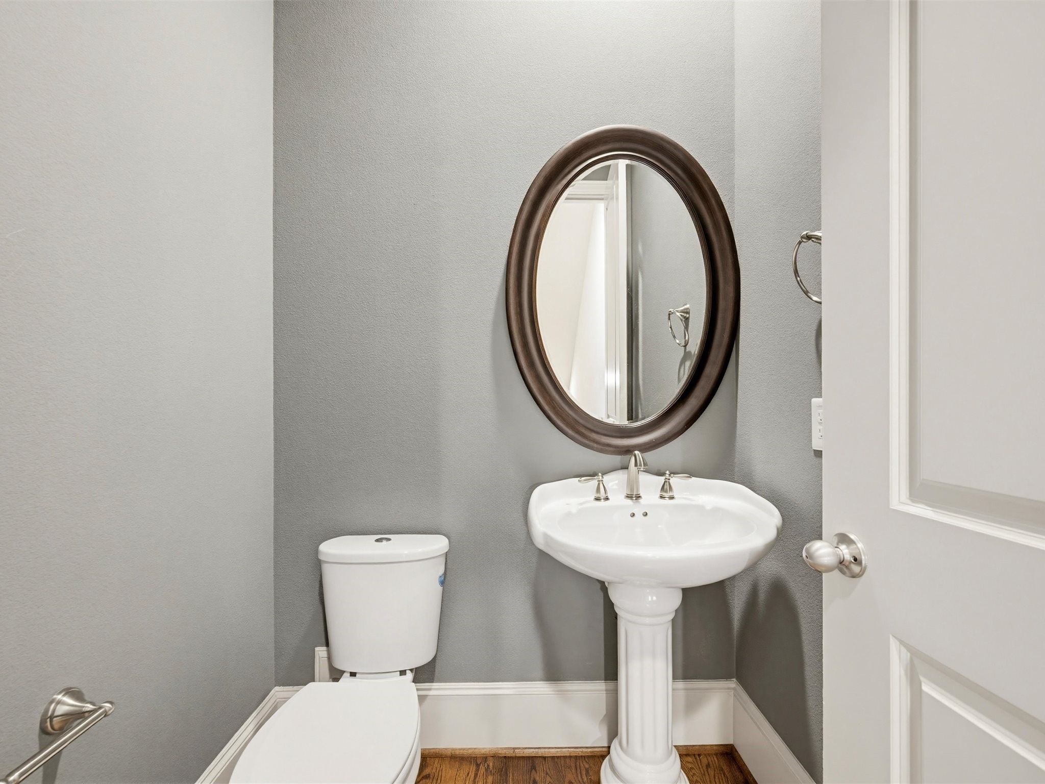 9427 London Bridge Station Houston, TX 77045 - Photo 15 of 30 This photo shows a small, elegant powder room featuring a pedestal sink with a round mirror above, a modern toilet, and gray walls complemented by wood flooring.