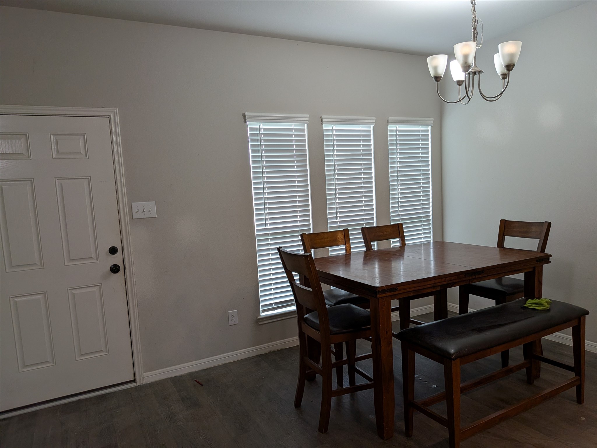 15730 Joe Dimaggio Street Splendora, TX 77372 - Photo 6 of 20 Cozy dining area featuring a wooden table with chairs and a bench, illuminated by a stylish chandelier. Natural light flows through three windows with blinds, complementing the neutral wall tones and wood flooring.