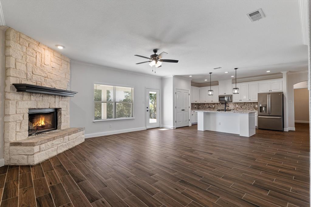3511 West J E Woody Road Springtown, TX 76082 - Photo 7 of 34 Unfurnished living room with crown molding, a stone fireplace, a ceiling fan, dark wood-style flooring, and recessed lighting