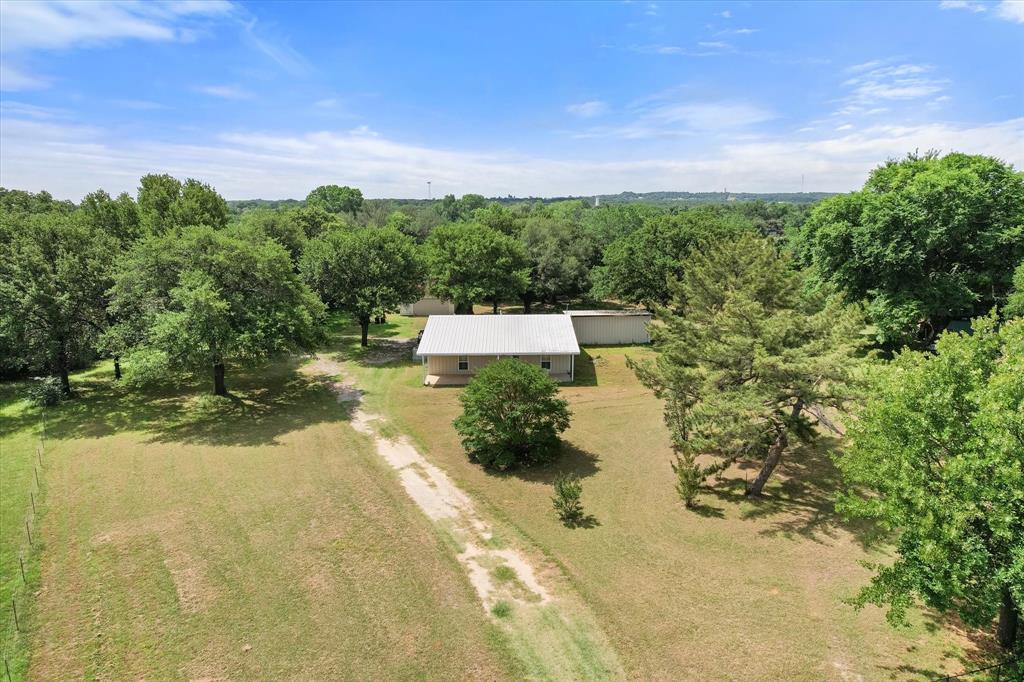 126 Smallwood Court Springtown, TX 76082 - Photo 22 of 24 Single-story residence with a metal roof surrounded by mature trees, featuring an extended gravel driveway and expansive grassy grounds