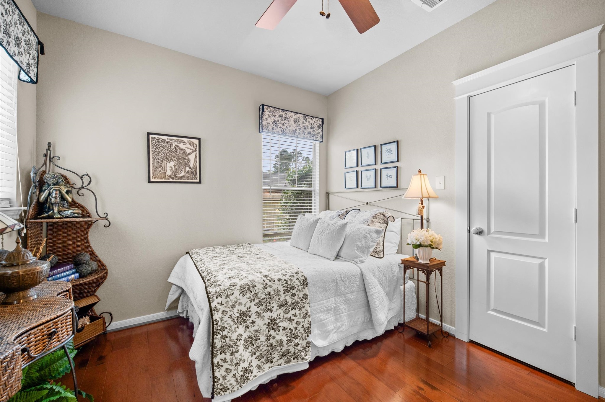 1603 Claytons Bend Court Spring, TX 77386 - Photo 12 of 38 Cozy bedroom with a neutral color palette, featuring a comfortable bed with floral bedding, hardwood floors, and ample natural light from two windows. Includes decorative wall art, a wicker shelf, and a bedside table with a lamp.