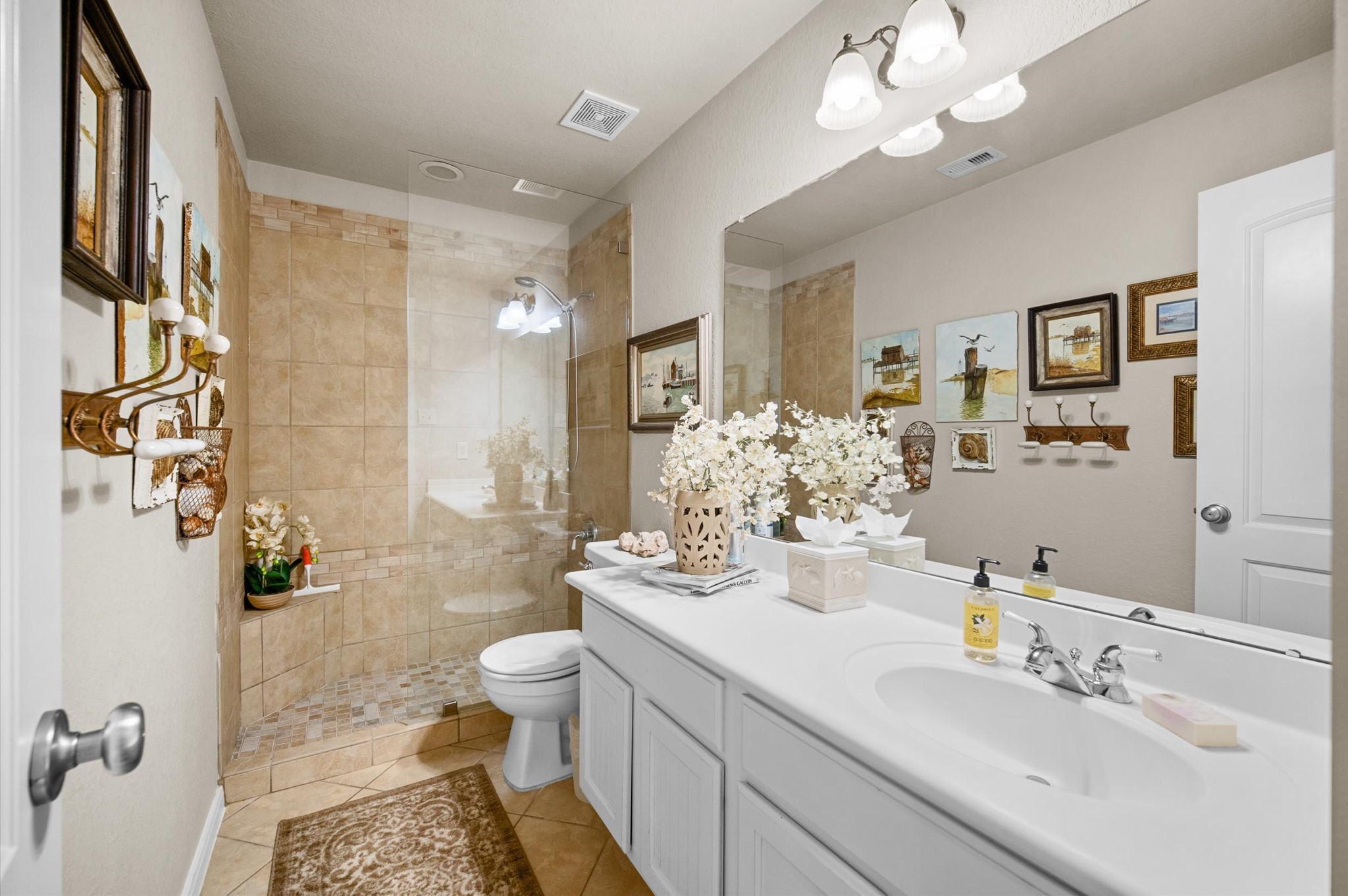 1603 Claytons Bend Court Spring, TX 77386 - Photo 10 of 38 Bright and elegant bathroom featuring a large mirror, white vanity with ample counter space, and a glass-enclosed shower with beige tiles. The decor includes floral arrangements and tasteful wall art, creating a welcoming atmosphere.