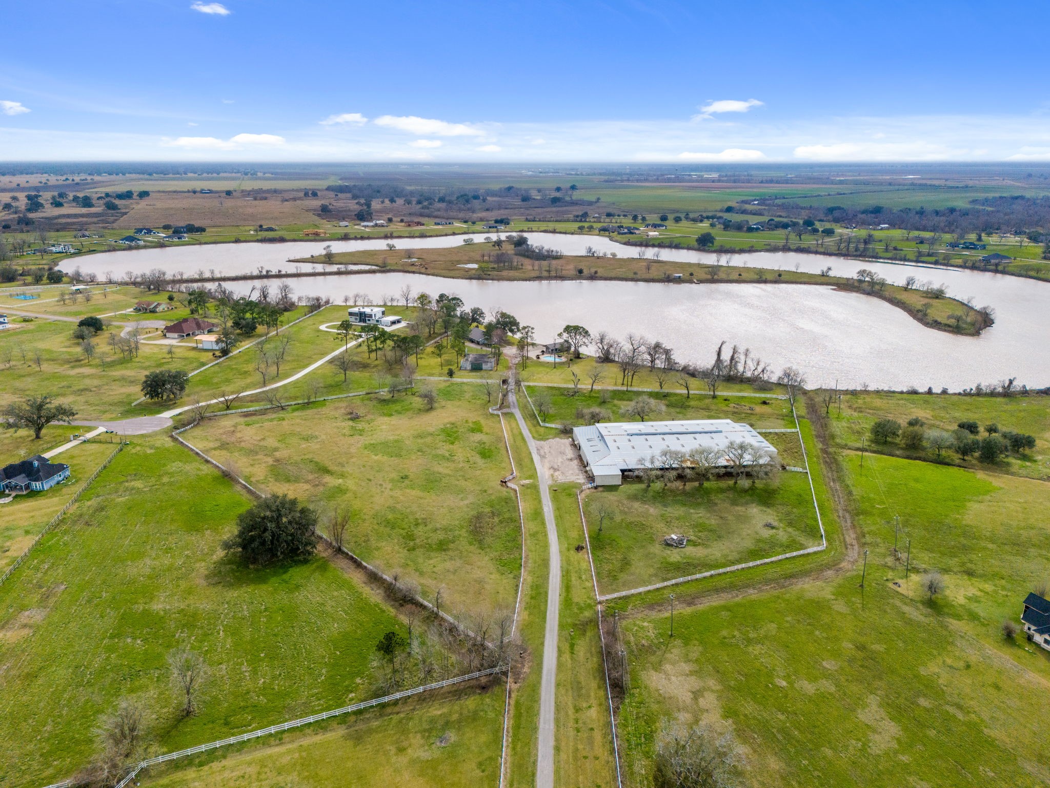 The lake stretches across the background, reflecting the expansive sky and reinforcing the property’s peaceful, resort-like feel. Wide lawns, scattered trees, and neighboring estates complete the scene, highlighting both the scale and versatility of this ranch setting.