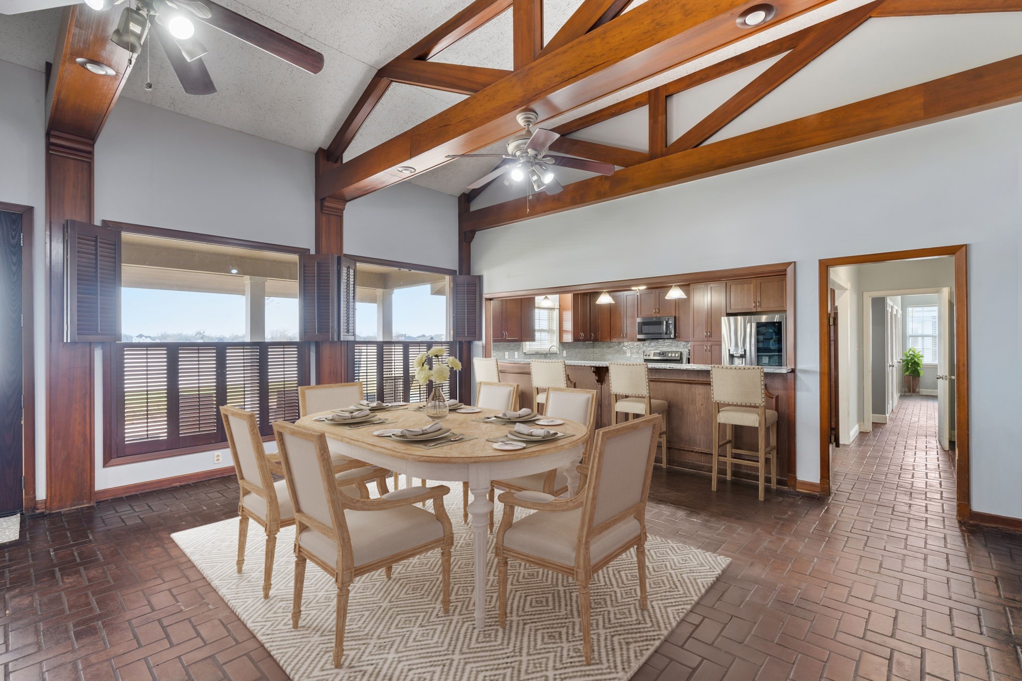 2935 West Fm 1462 Rosharon, TX 77583 - Photo 14 of 50 Virtually Staged: This open-concept ranch dining space showcases classic craftsmanship and a warm, inviting atmosphere. Soaring ceilings with exposed wood trusses and beams create a dramatic yet grounded ranch aesthetic, while ceiling fans and abundant natural light add everyday comfort. The dining area is perfectly positioned for gatherings, anchored by a round table that encourages conversation and easy flow. Brick flooring adds durability and timeless character, ideal for ranch living.