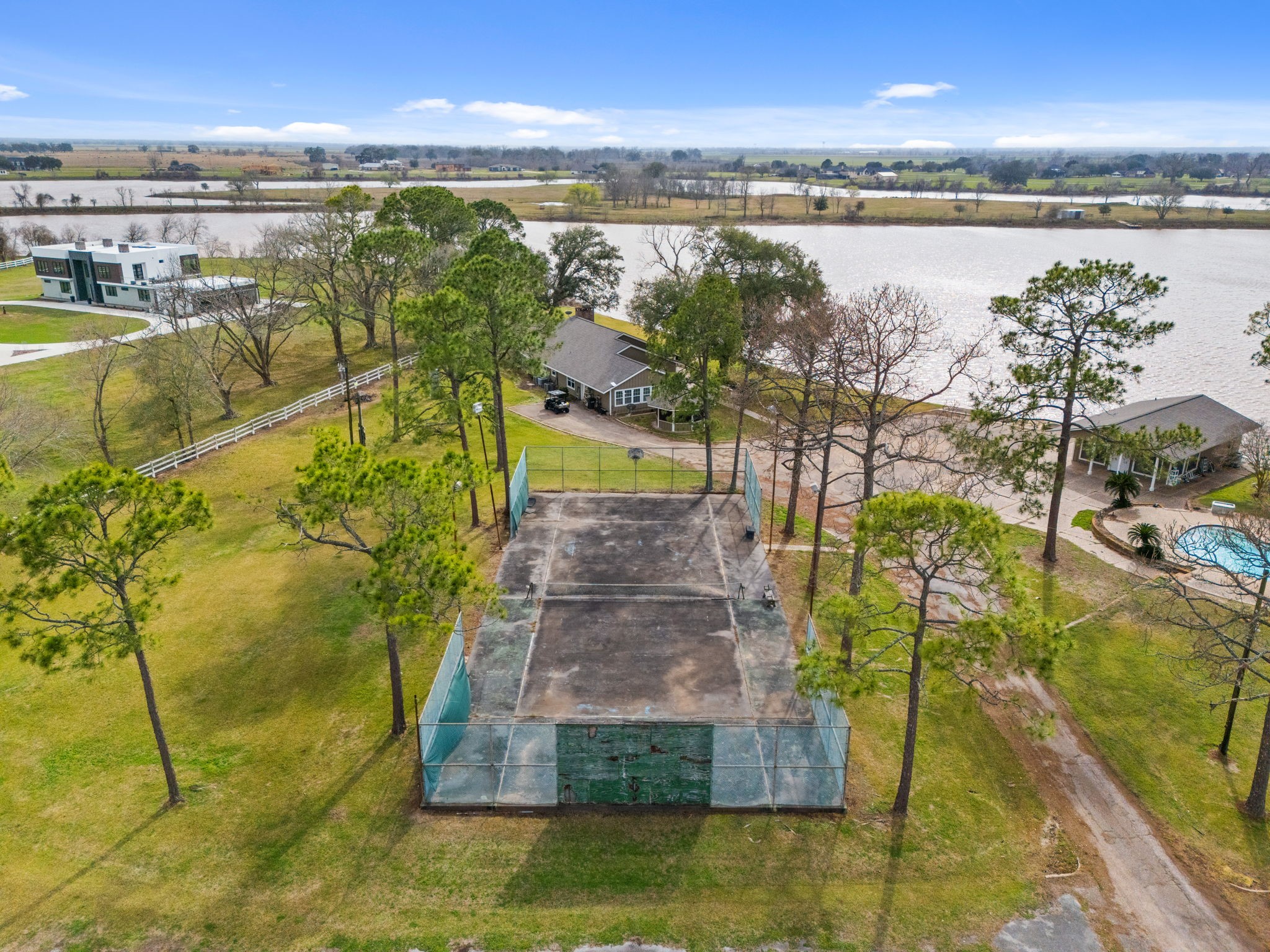 2935 West Fm 1462 Rosharon, TX 77583 - Photo 47 of 50 This elevated aerial view captures a unique mix of recreation, waterfront living, and open ranch space. Centered in the frame is a fenced tennis court nestled among mature pine trees, offering privacy and a shaded setting.