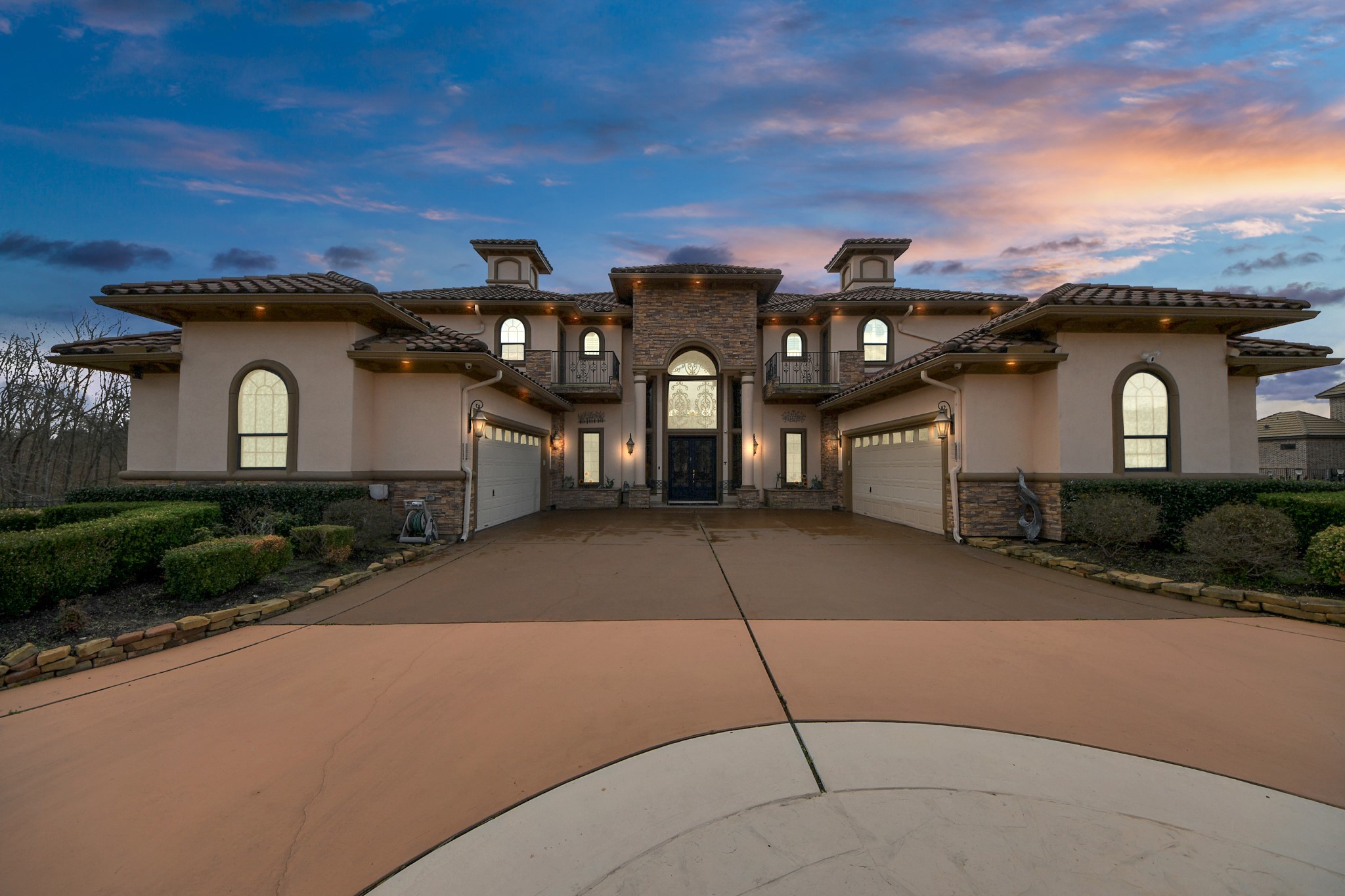 This stunning two-story home features a grand entrance with a stone façade, arched windows, and a four-car garage. The manicured landscaping and expansive driveway enhance its curb appeal, set against a picturesque sunset backdrop.