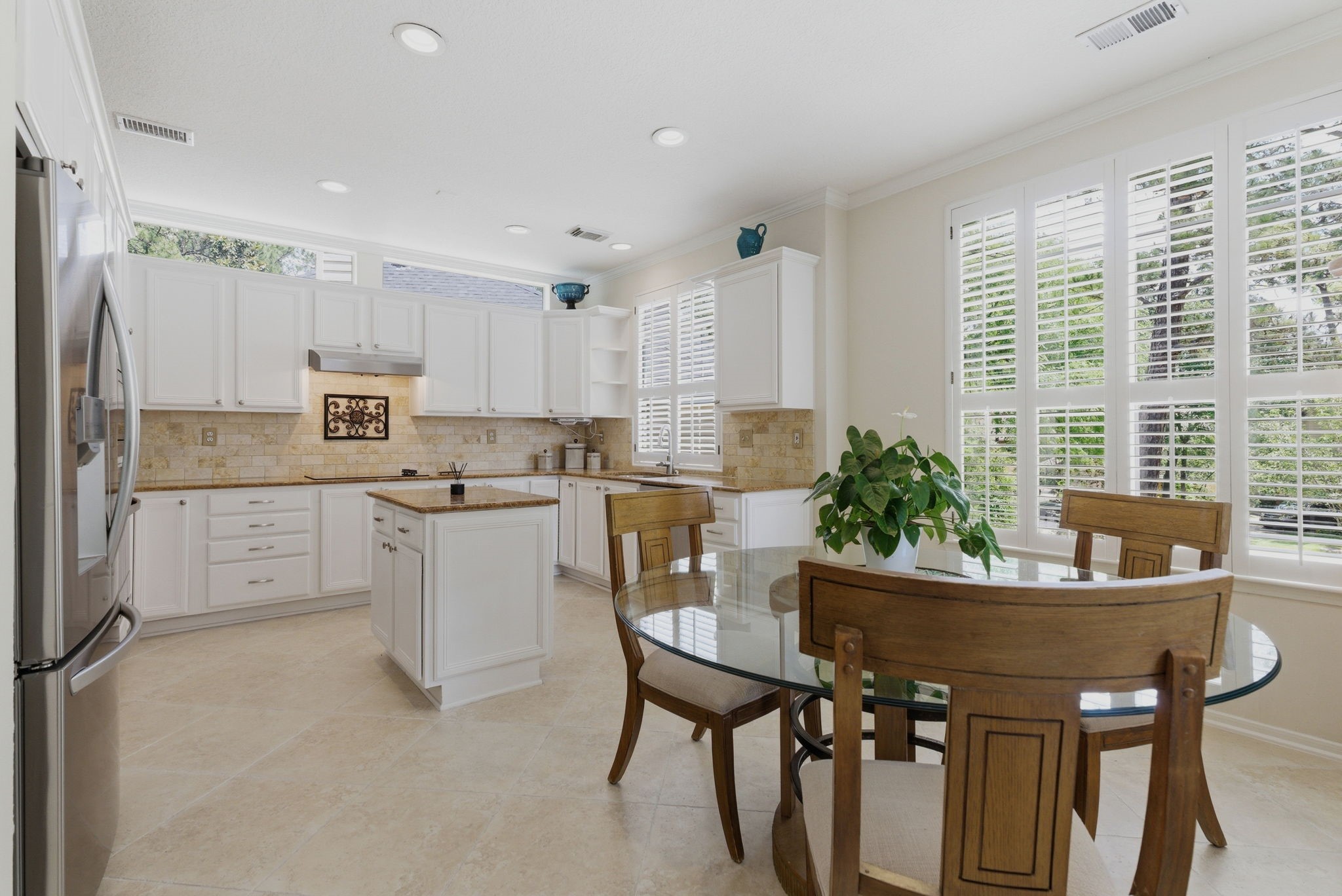 11 Dovewing Place Spring, TX 77382 - Photo 9 of 32 Bright, spacious kitchen with white cabinetry, stainless steel appliances, and a central island. Features a cozy dining area with a glass table and wooden chairs, complemented by large windows with shutters providing ample natural light.