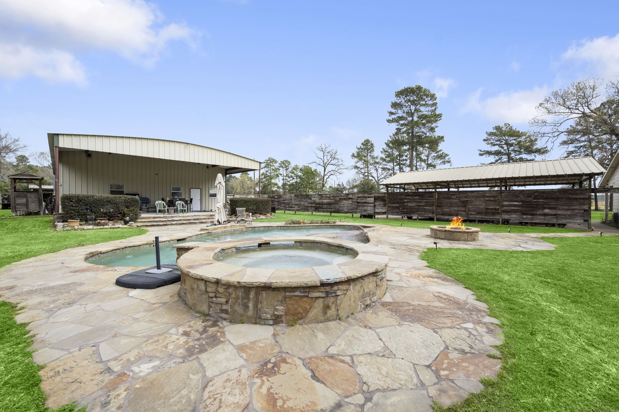 26136 Joseph Road Hockley, TX 77447 - Photo 17 of 31 This backyard features a spacious patio with a stone hot tub, a fire pit, and ample seating. The area is surrounded by lush greenery and includes a covered outdoor space, perfect for entertaining or relaxing.