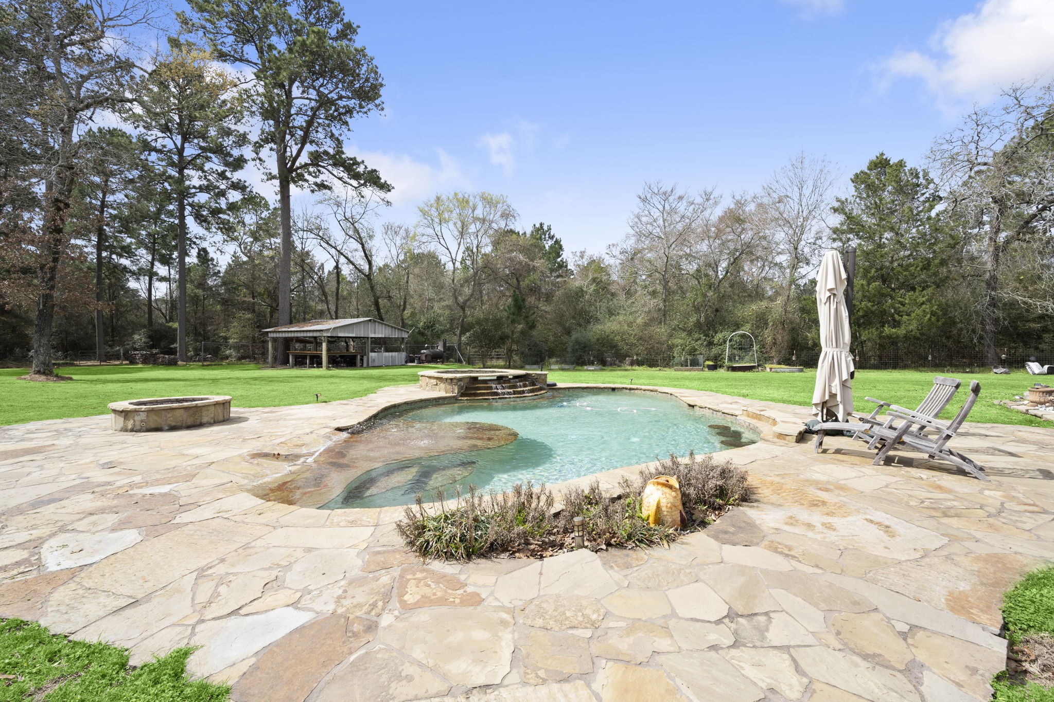 26136 Joseph Road Hockley, TX 77447 - Photo 18 of 31 This photo showcases a spacious backyard featuring a natural stone patio with a swimming pool. The pool area is surrounded by lush greenery and mature trees, offering a serene and private outdoor retreat. There are lounge chairs and an umbrella for relaxation, and a separate structure in the background adds functionality.