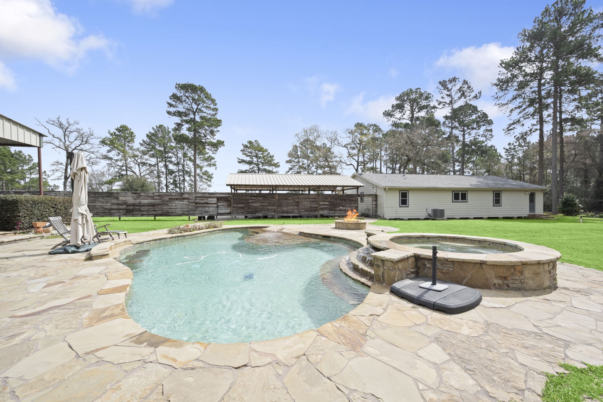 26136 Joseph Road Hockley, TX 77447 - Photo 19 of 31 This photo showcases a spacious backyard featuring a stone patio with an inviting pool and attached hot tub. A fire pit adds warmth and ambiance, and the area is surrounded by lush greenery and tall trees, offering a serene and private outdoor space.