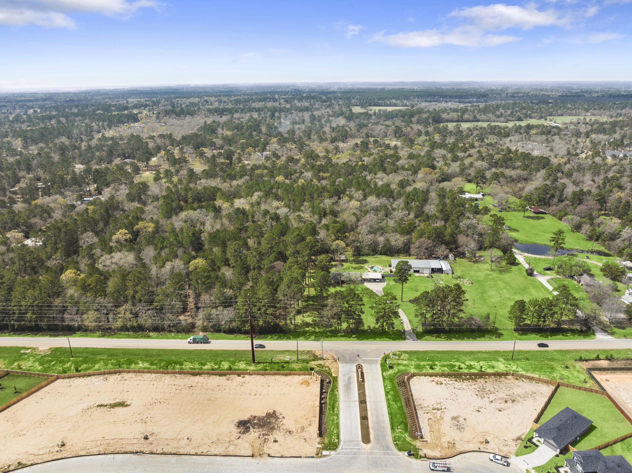 26136 Joseph Road Hockley, TX 77447 - Photo 2 of 31 This aerial photo shows a scenic, wooded area with vast greenery, a home, building, and a small pond. The foreground features cleared land, potentially for new development, bordered by a road. Ideal for those seeking tranquility and space.