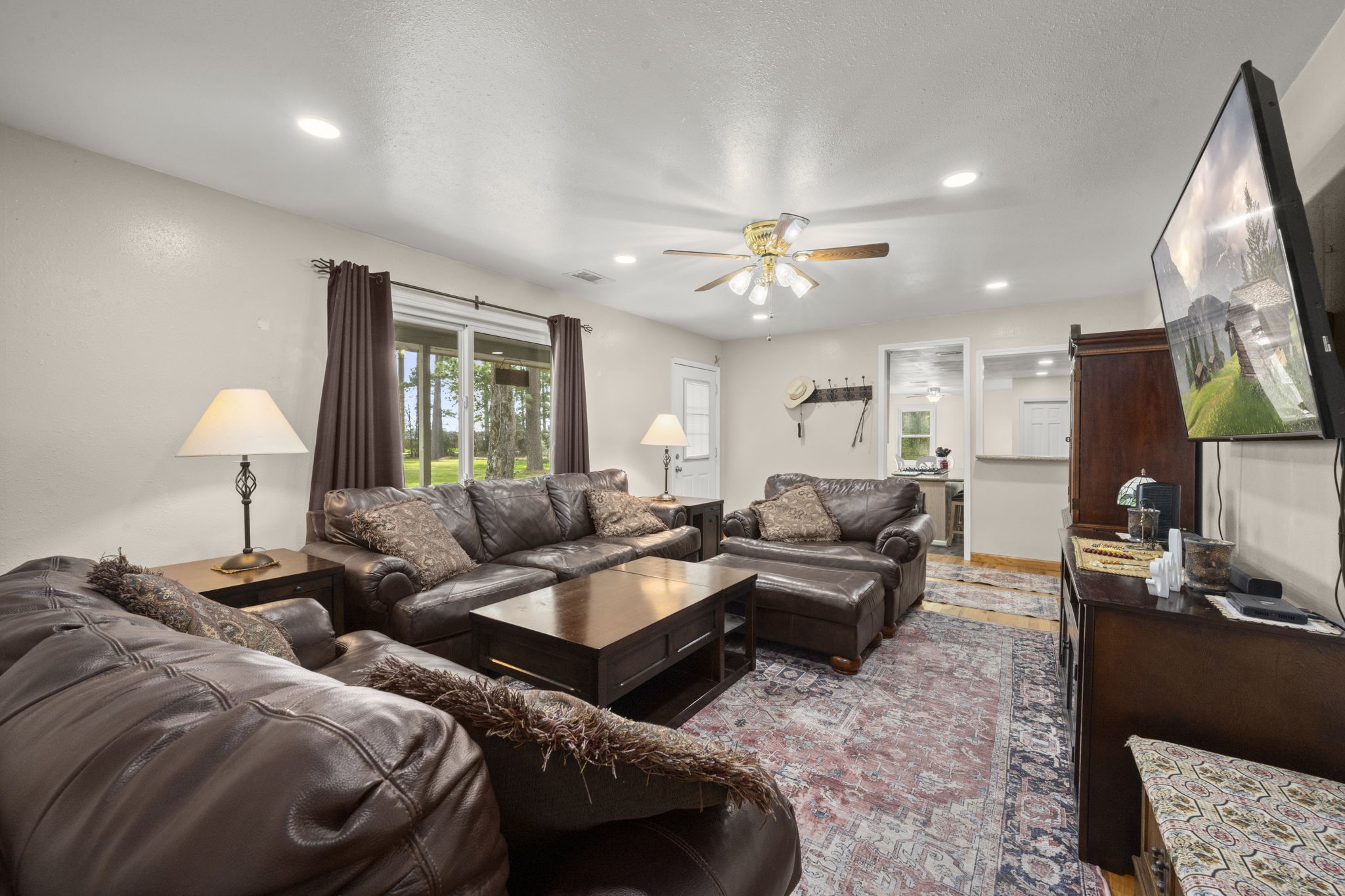 26136 Joseph Road Hockley, TX 77447 - Photo 21 of 31 Spacious living room featuring comfortable leather sofas, a ceiling fan, large window with natural light, and a mounted TV. The room has a cozy, inviting atmosphere with warm lamps and a decorative rug. It opens to other areas, offering a seamless flow.