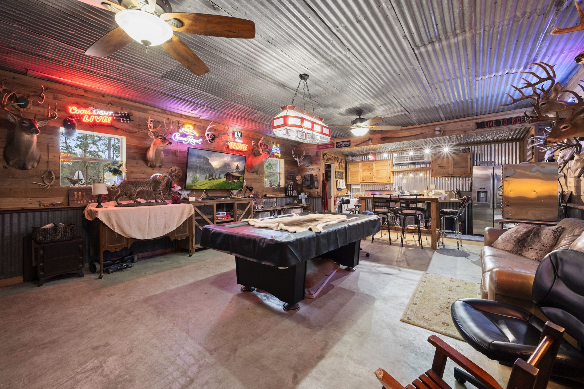 26136 Joseph Road Hockley, TX 77447 - Photo 26 of 31 Metal building/shop has a rustic game room with a corrugated metal ceiling and wood-paneled walls. It features a pool table, mounted animal trophies, neon signs, and a bar area with counter seating. The space is cozy and inviting, ideal for entertainment and relaxation.