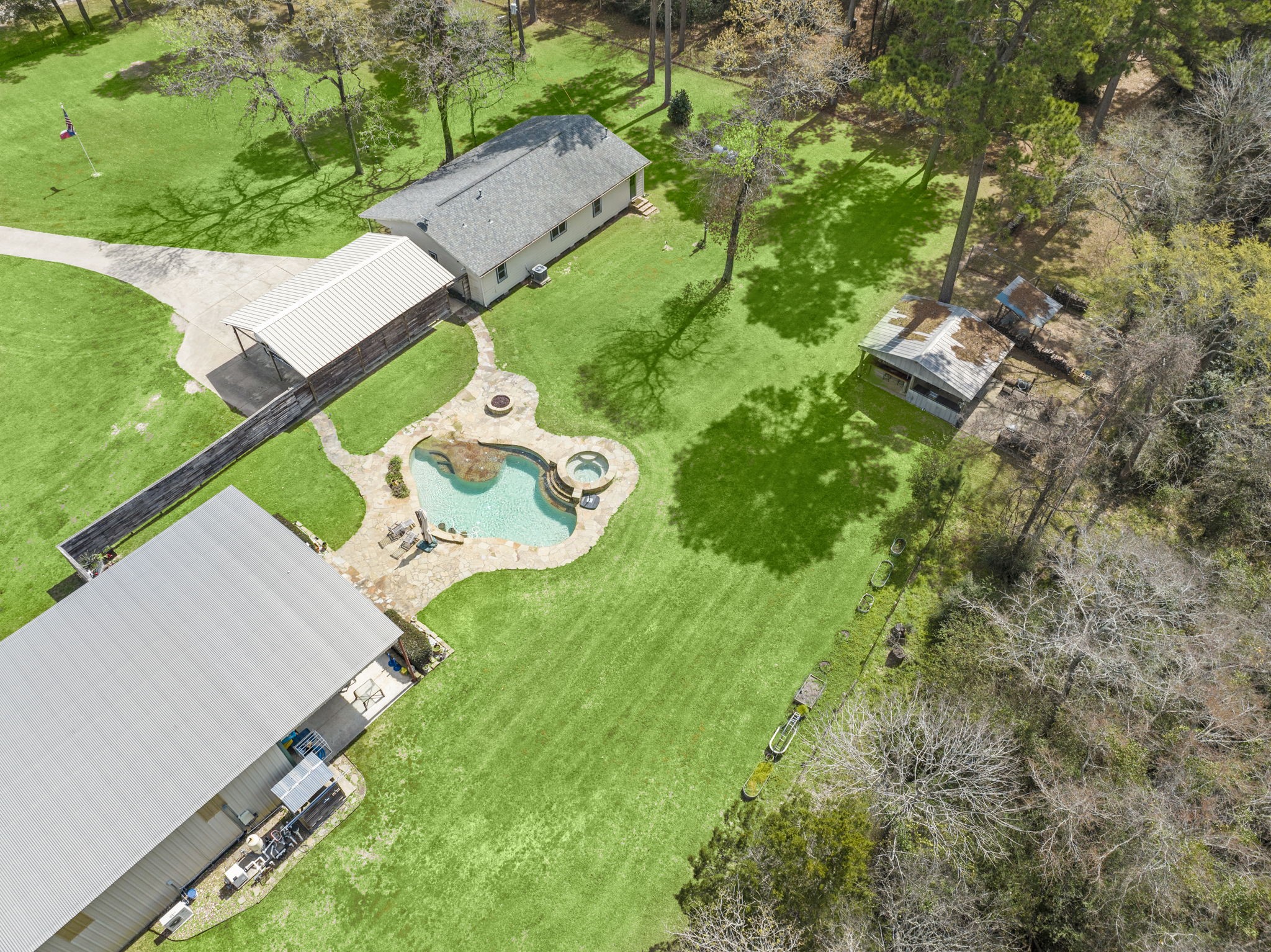 26136 Joseph Road Hockley, TX 77447 - Photo 9 of 31 This aerial view showcases a spacious property with a large, well-maintained lawn, a swimming pool with a spa, and multiple outbuildings. Tall trees provide shade, and there's a long driveway leading to a carport. Ideal for privacy and outdoor living.