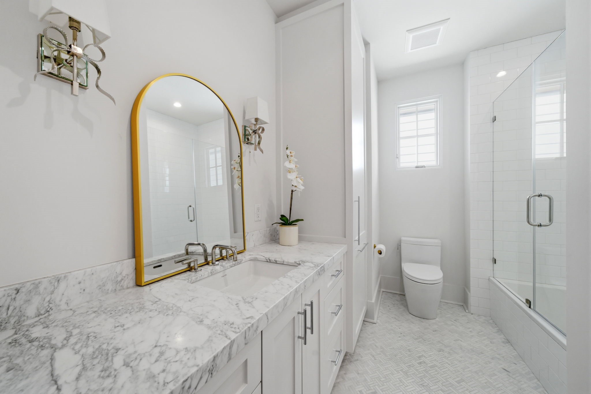 2615 Yupon Street Houston, TX 77006 - Photo 38 of 50 Spa-inspired primary ensuite bathroom features an expansive white custom vanity topped with a thick marble counter and a modern trough-style sink. A gold-framed arch mirror and ribbon-accented sconces add a touch of luxury, while the large-format stone tile flooring flows toward a glass-enclosed shower and soaking tub. Natural light from a transom window enhances the bright, airy atmosphere of this contemporary 2022 custom estate retreat.
