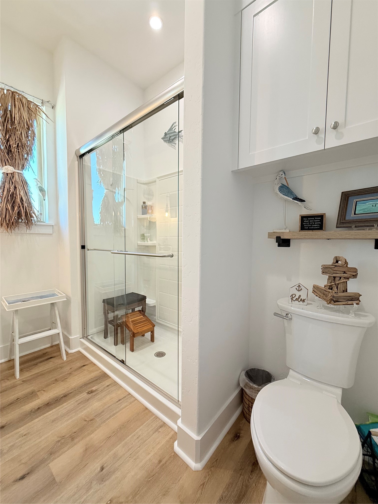 2023 County Road 201 Sargent, TX 77414 - Photo 29 of 45 This is a bright, modern bathroom featuring a glass-enclosed shower, white walls, and light wood flooring. It has a cozy, decorative touch with shelving above the toilet and a small window for natural light.