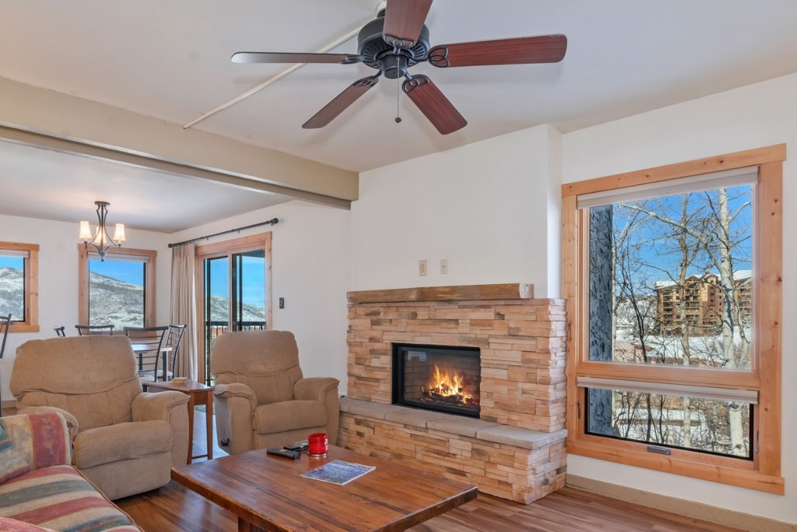 Living room with beam ceiling, a stone fireplace, wood finished floors, and ceiling fan with notable chandelier