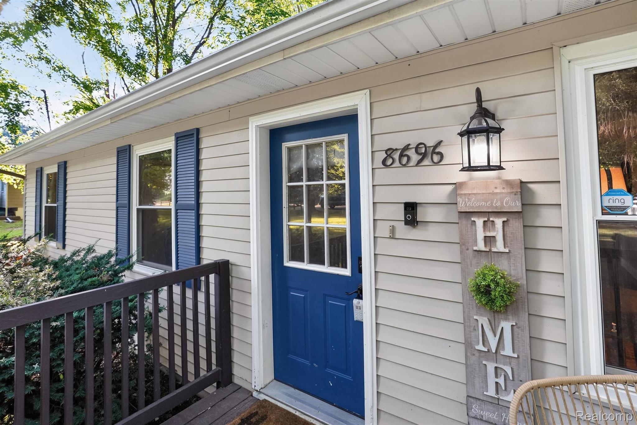 8696 West Sargent Road Fowlerville, MI 48836 - Photo 2 of 36 Exterior siding features a blue paneled entry door with sidelights, contrasting blue window shutters, and a dark wood-finish railing