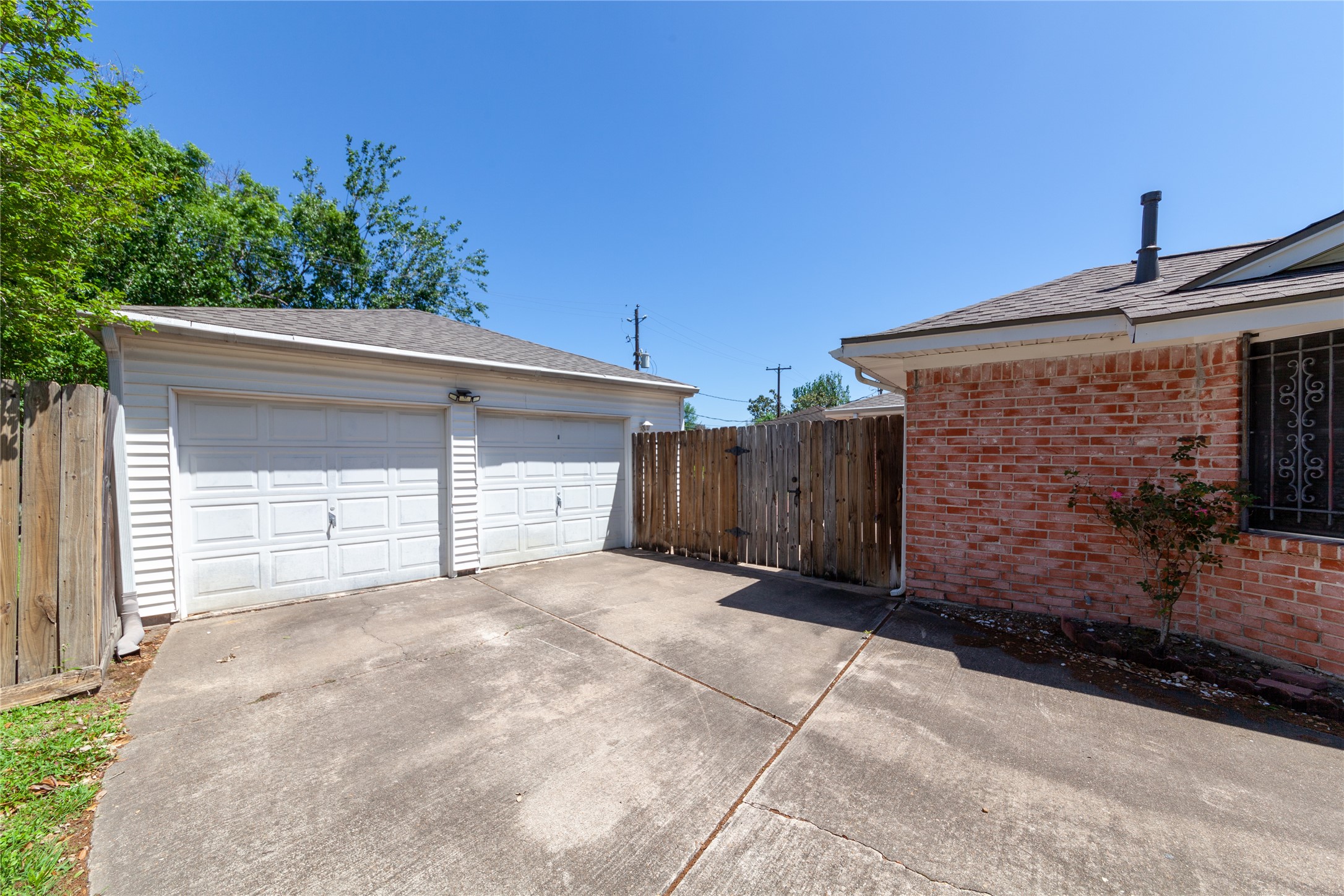 8826 Roos Road Houston, TX 77036 - Photo 37 of 42 A spacious driveway leading to a two-car garage with white doors. A brick exterior wall and wooden fence offer privacy, while the area is surrounded by greenery, providing a well-maintained and inviting outdoor space.