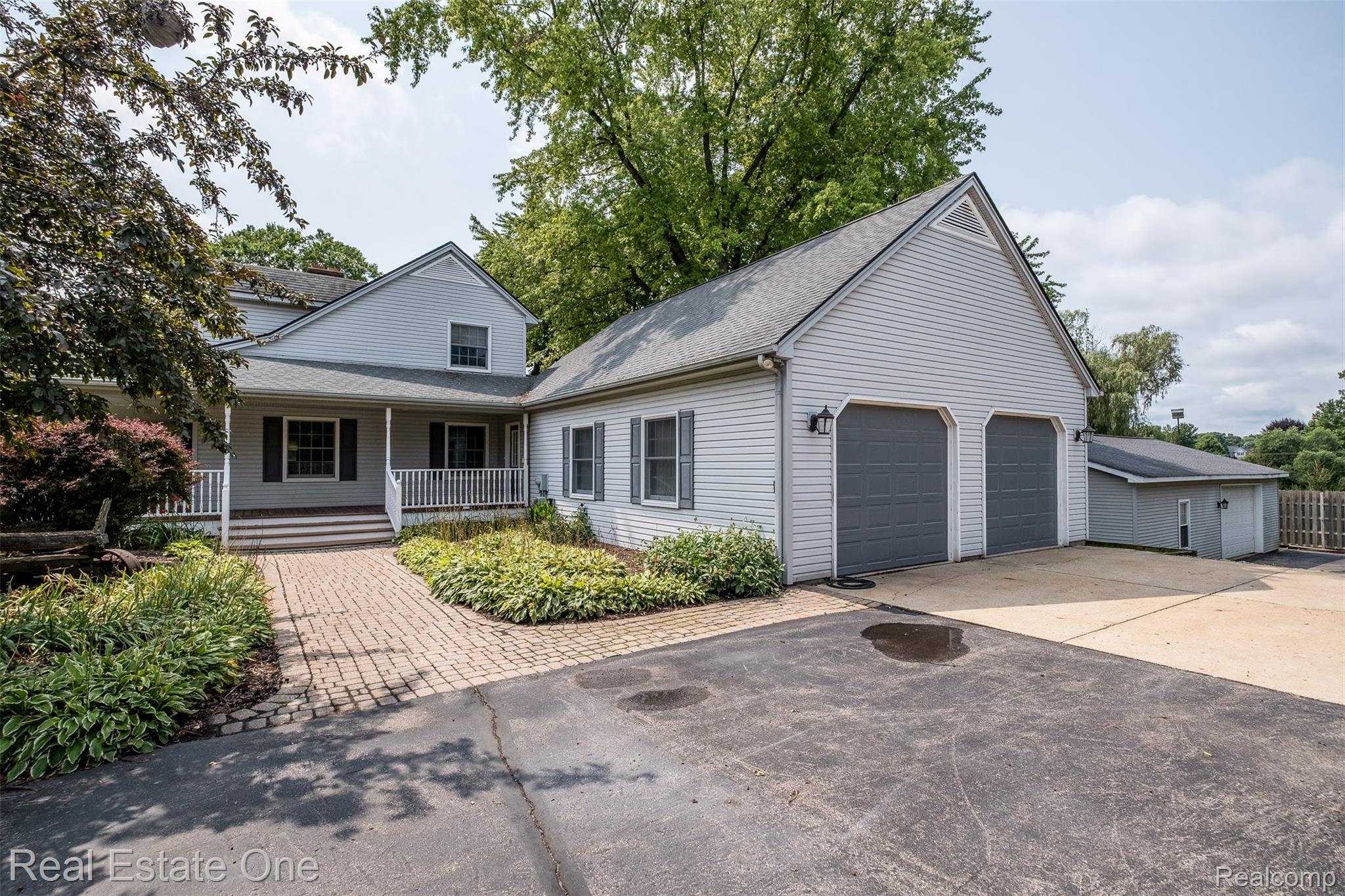 11030 Marshall Road South Lyon, MI 48178 - Photo 63 of 83 Contemporary home featuring a brick paver driveway, two-car attached garage with dark panel doors, vinyl siding exterior, covered front porch with railings, and mature landscaping with established trees throughout the property