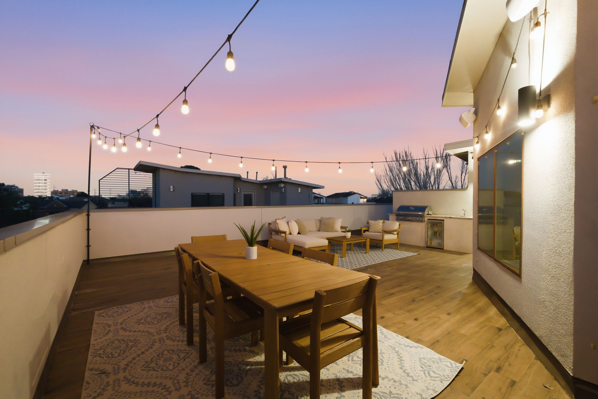 1405 Vermont Street, Unit A Houston, TX 77006 - Photo 40 of 46 Glow of string lights illuminates this expansive rooftop deck, creating a sophisticated setting for evening gatherings. Low-maintenance wood-look flooring leads to a high-end summer kitchen, while the spacious layout comfortably accommodates a plush lounge area for enjoying sunset views over the city. A designer geometric rug and sleek furniture enhance the luxury contemporary atmosphere of this private Bradford Mews retreat.