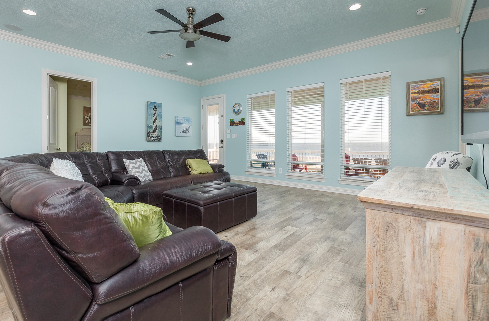 843 South Jacks Road Crystal Beach, TX 77650 - Photo 23 of 45 Spacious and bright living room with large windows, light blue walls, and gorgeous flooring. Features a comfortable leather sectional sofa, ceiling fan, and a coastal wooden TV stand. Ideal for relaxing and entertaining.
