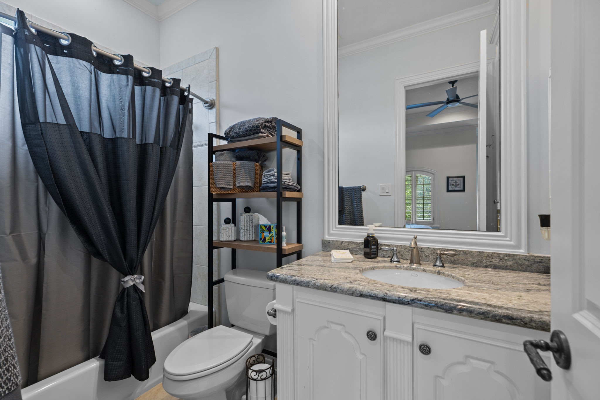 39 Maymont Way Spring, TX 77382 - Photo 31 of 50 This bathroom features a sleek design with a granite countertop, white cabinetry, and modern fixtures. It includes a shower with a dark curtain, a toilet, and a shelving unit for storage. A large mirror adds depth, and there's ample lighting.