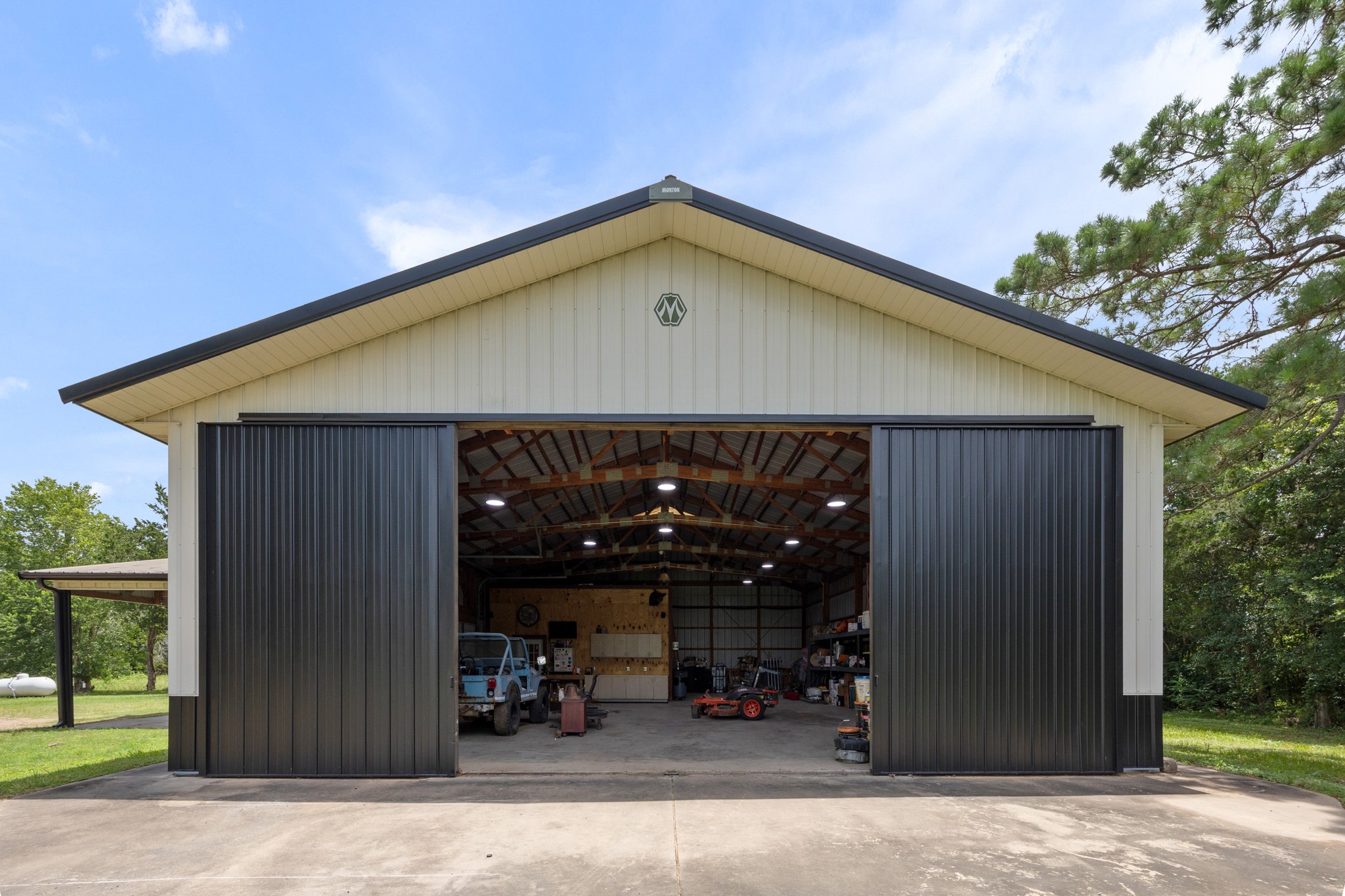 10209 Oberrender Road Needville, TX 77461 - Photo 40 of 50 This photo shows the spacious 40X60 metal workshop with large sliding doors, ample interior space for vehicles or equipment, and a sturdy overhang. It sits on a concrete pad, surrounded by greenery, offering a versatile addition to any home.