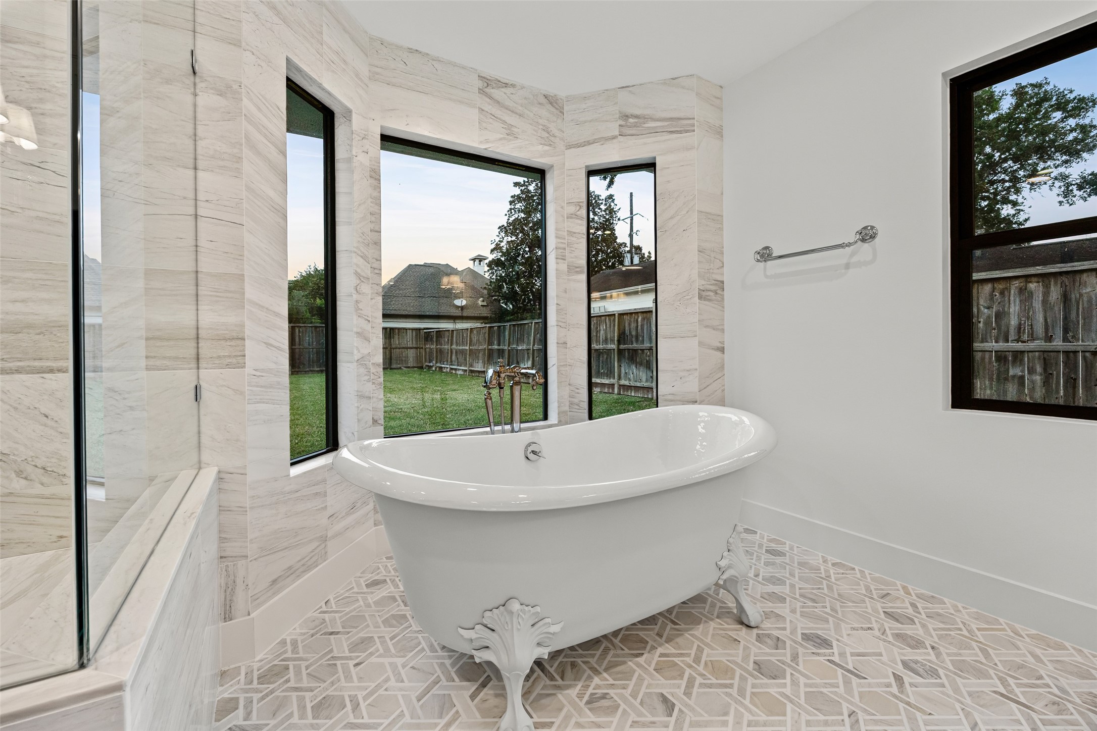5658 Del Monte Drive Houston, TX 77056 - Photo 19 of 35 The spa bathroom is a masterpiece of craftsmanship and luxury. Wrapped in stunning full-height marble-look tile, the walk-in shower offers dual showerheads, a built-in bench, and a large niche with hexagon mosaic detailing. A freestanding clawfoot tub sits perfectly in front of floor-to-ceiling picture windows, offering serene backyard views and natural light that elevates the entire space.