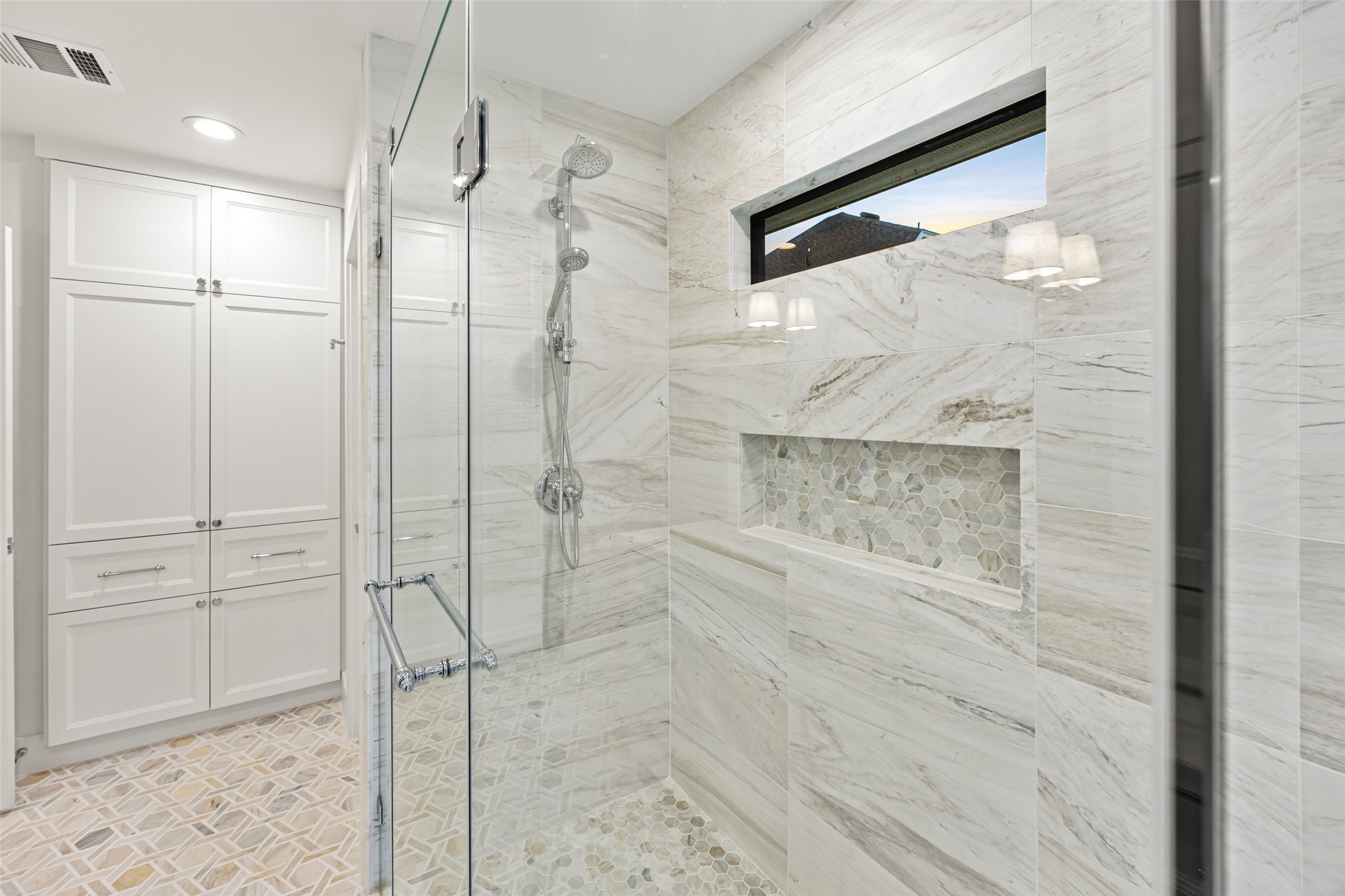 5658 Del Monte Drive Houston, TX 77056 - Photo 22 of 35 Step into a beautifully curated primary shower that blends timeless elegance with modern luxury. Wrapped in dramatic large-format marble-look tile, this space delivers a serene, spa-level experience from the moment you enter. A frameless glass enclosure showcases the exquisite design, while the oversized hexagon mosaic niche adds both functionality and visual texture.
Dual showerheads—including a rainfall feature and handheld wand—provide elevated comfort, and a sleek built-in bench creates the perfect s