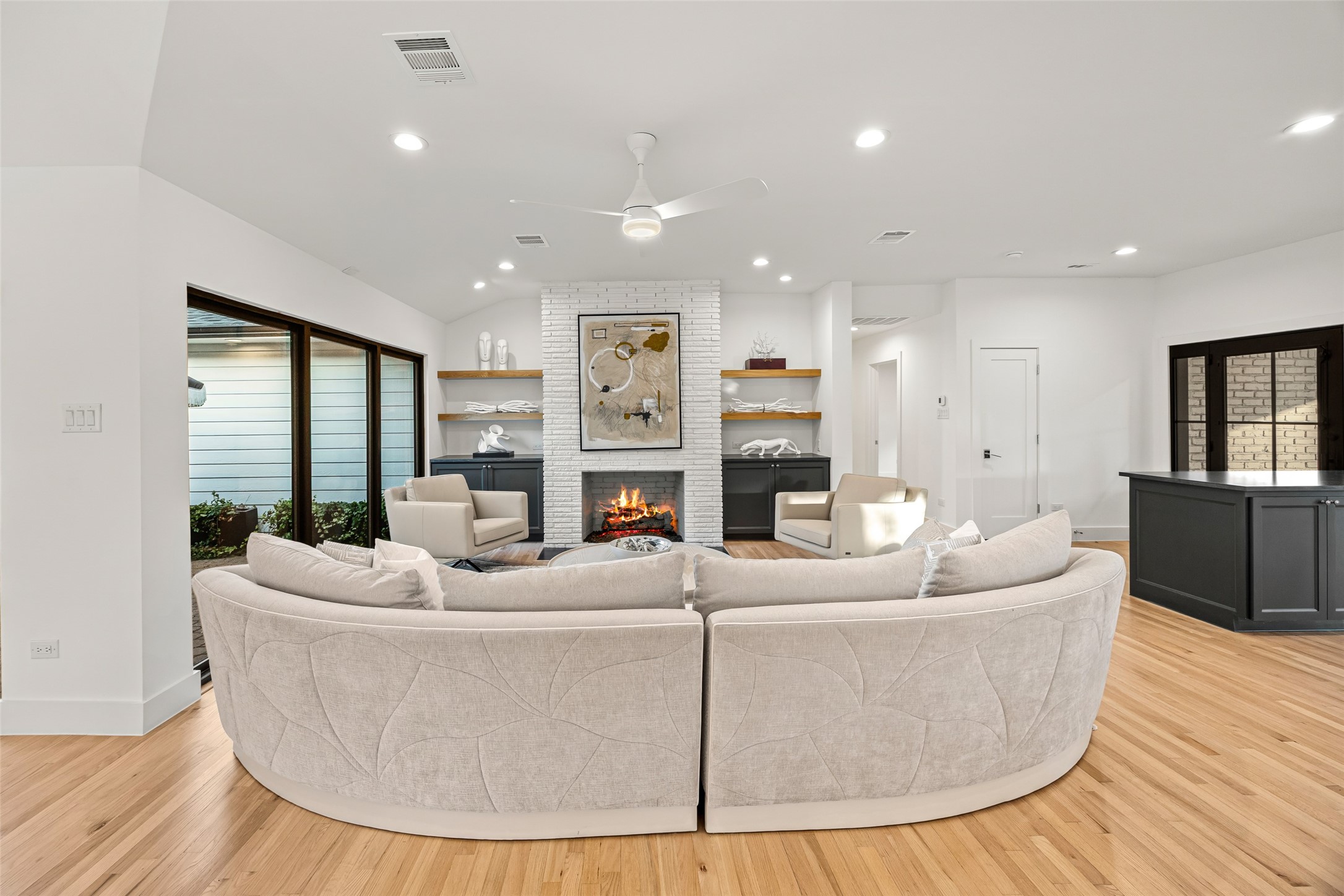 5658 Del Monte Drive Houston, TX 77056 - Photo 27 of 35 Welcome to a designer-crafted, fully reimagined single-story home that truly defines modern luxury living. Flooded with natural light and wrapped in floor-to-ceiling windows, this expansive open-concept living space seamlessly connects the formal dining room, living room, and chef’s kitchen—creating a rare, effortless flow ideal for both daily living and elevated entertaining.
The living room centers around a custom white-brick fireplace framed by bespoke cabinetry and floating wood shelves, adding archi