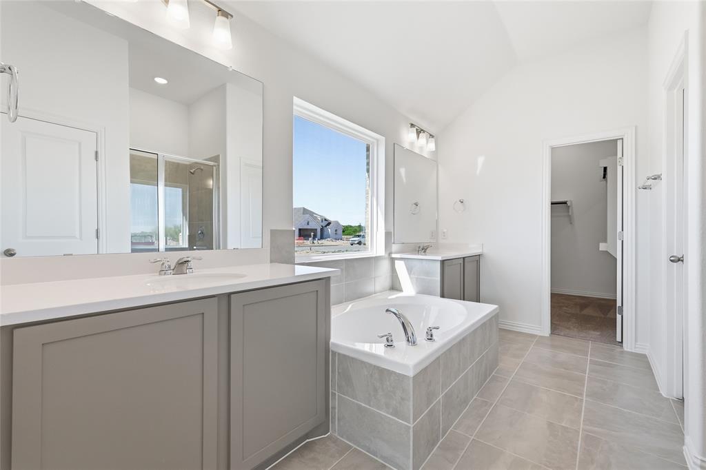 2529 Rivers W Road Anna, TX 75409 - Photo 23 of 40 Spacious bathroom featuring a built-in tub with tiled surround, dual vanities with white countertops and undermount sinks, a large window, a separate shower with glass enclosure, and vaulted ceilings
