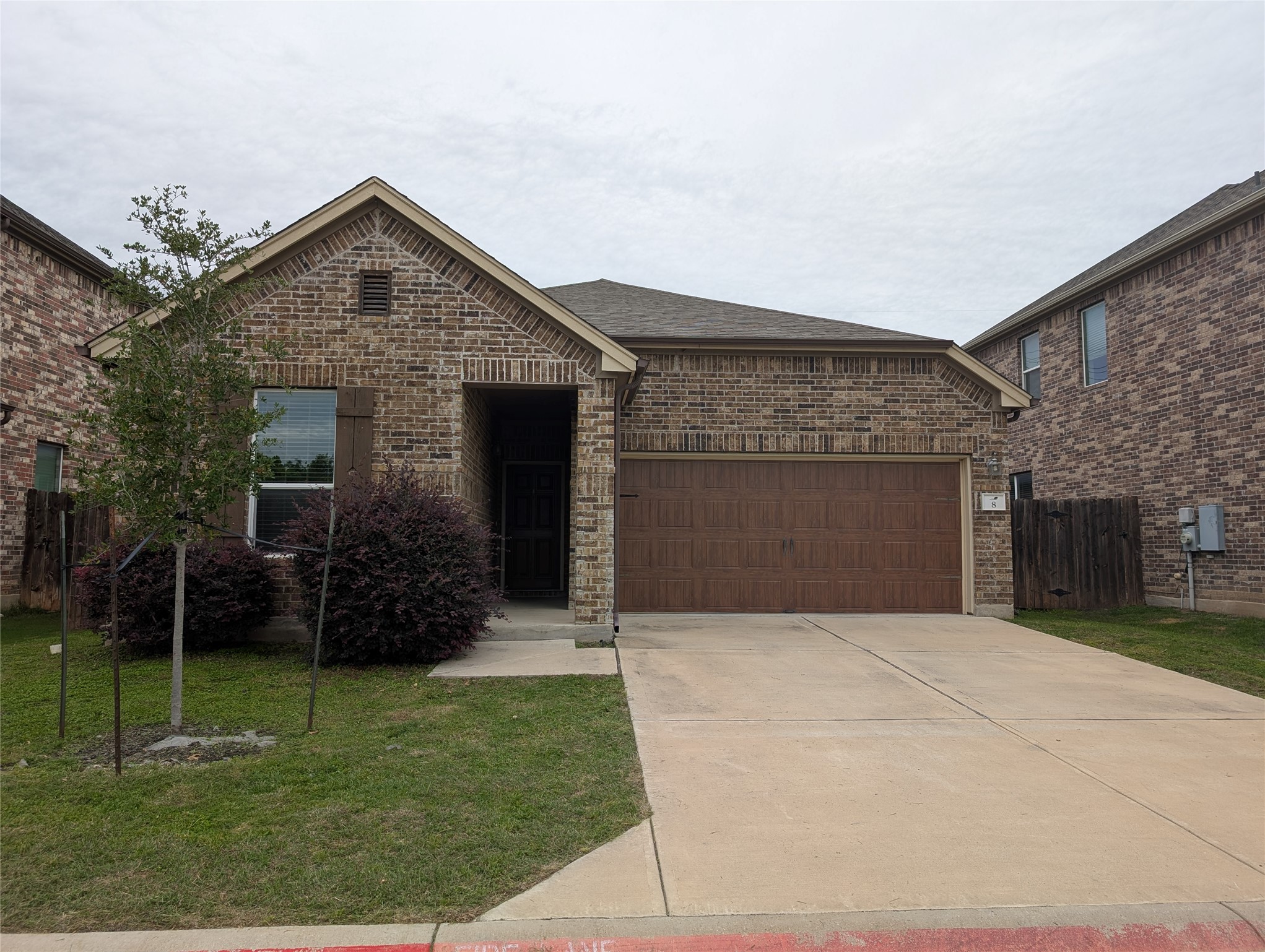 Brick exterior with a peaked gable, a two-car garage with a brown door, a paved driveway, and mature shrubs