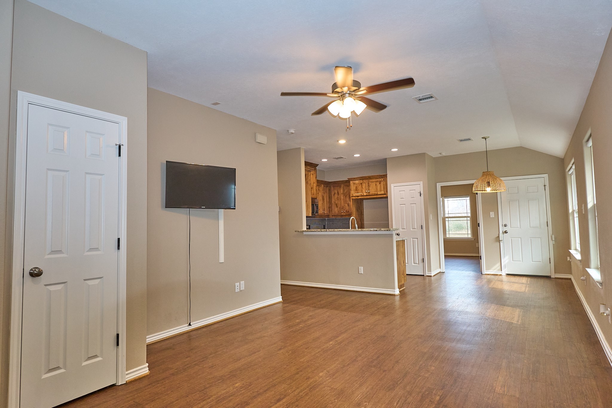 1807 Nuches Lane Bryan, TX 77803 - Photo 2 of 27 This photo shows an open-concept living space with wood flooring and neutral walls. It features a ceiling fan, mounted TV, and a kitchen with wooden cabinets and a breakfast bar. Natural light streams in through several windows, creating a bright and inviting atmosphere.