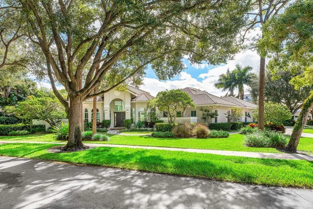 a view of a white house in front of a big yard with plants and large trees
