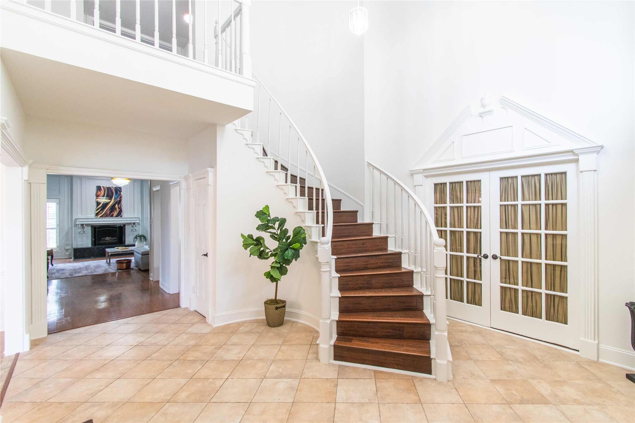 Spacious foyer features elegant tiled flooring and a striking curved wooden staircase with white railings. French doors add a touch of sophistication, while the open layout leads to a cozy living area with a fireplace, creating a welcoming atmosphere.