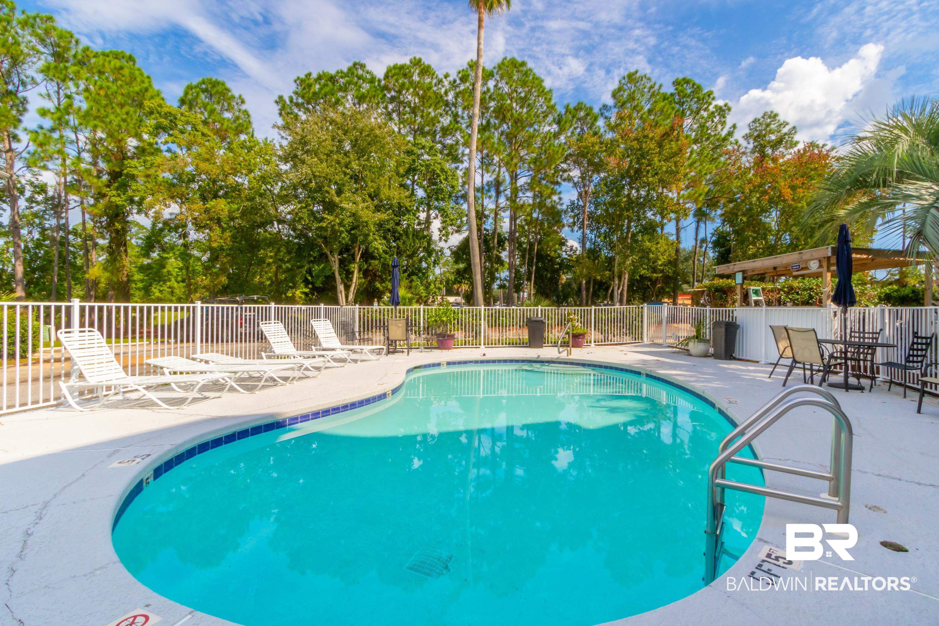 25957 Canal Road, Unit 208 Orange Beach, AL 36561 - Photo 26 of 28 Curved swimming pool featuring a white perimeter fence, surrounding mature trees, and a poolside pergola with built-in bar