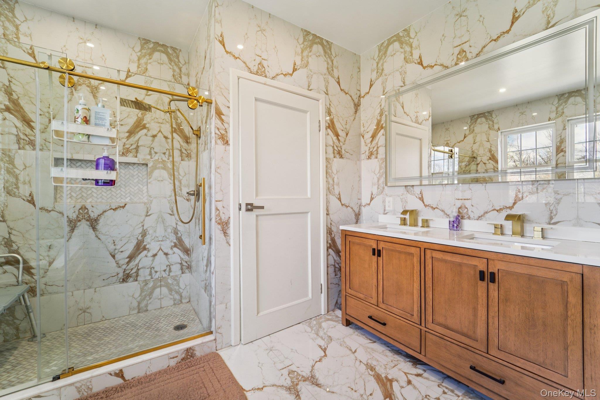 109 Bennett Road Newburgh, NY 12550 - Photo 26 of 41 This bathroom features elegant marble-patterned tile flooring and walls, a spacious shower with brass-toned fixtures and a glass enclosure, and a double vanity with a wooden cabinet and white countertops.