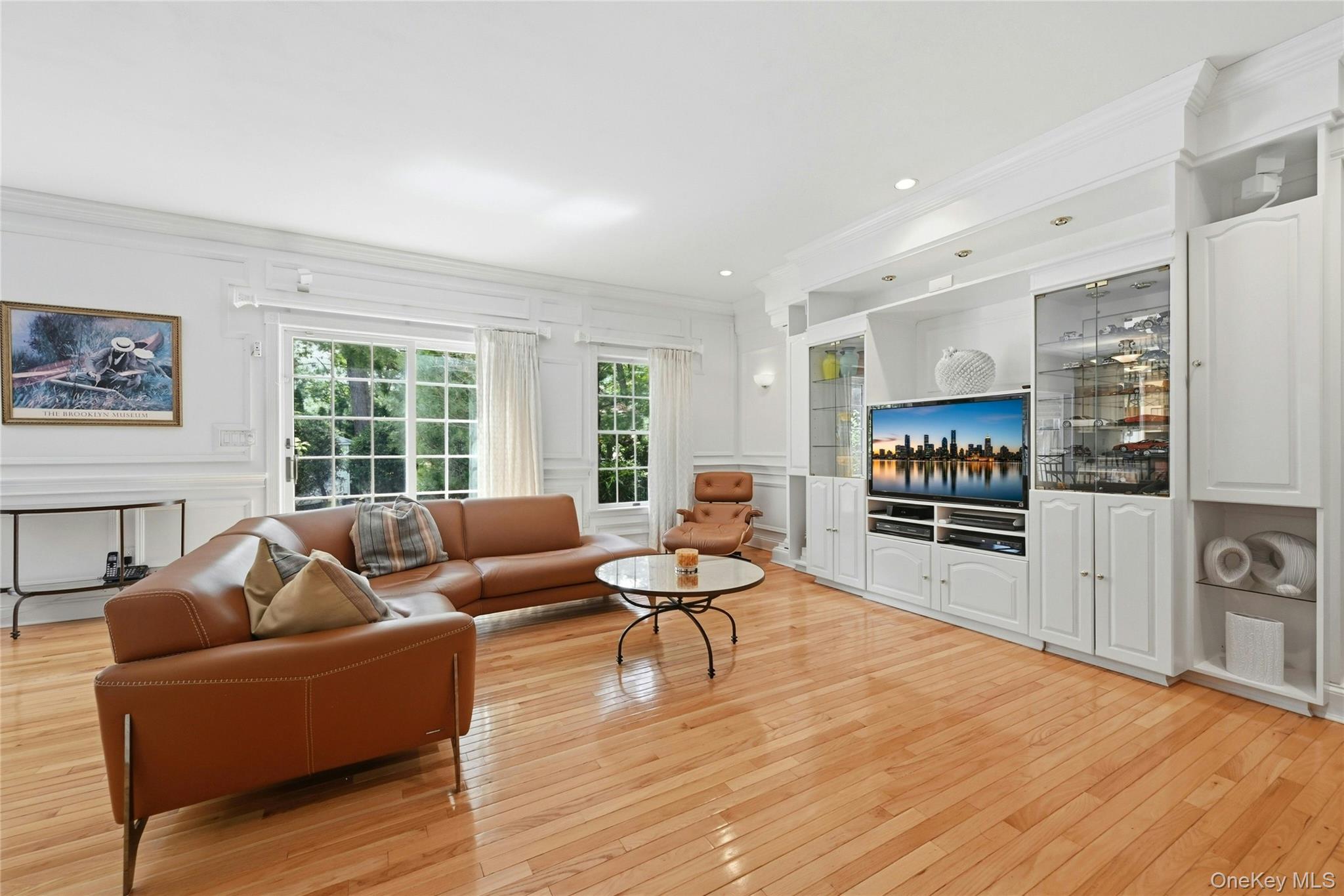 6 Thunder Road Miller Place, NY 11764 - Photo 21 of 50 The Expansive 20'3" x 18'8" Den/Family Room Was Expanded Two Feet Out The Back Of The Home & An Additional Two Feet Towards The Garage Offering A Perfect, Vast Space For Entertaining With 9' Ceilings, Stunning Box Paneled/Wainscoted Walls, Beautiful Hardwood Floors, Andersen Sliding Doors & Oversized Windows Overlooking The Backyard & In Ground Pool, A Striking Custom Built Wall Unit/Entertainment Center With Magnificent Crown Moldings, Columns, Glass Doors & Shelving, Built In Television (Which Stays With The Home) & Built In Lighting, Ample Room For A Very Large Furniture/Seating Area, And Access To The Half Bathroom/Powder Room & Two Car Garage.