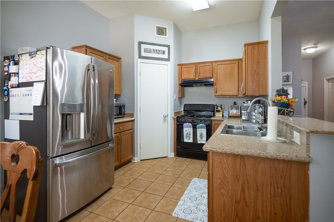 904 Crested Point Drive College Station, TX 77845 - Photo 5 of 11 Kitchen featuring appliances with stainless steel finishes, a peninsula, brown cabinetry, light tile patterned flooring, and under cabinet range hood