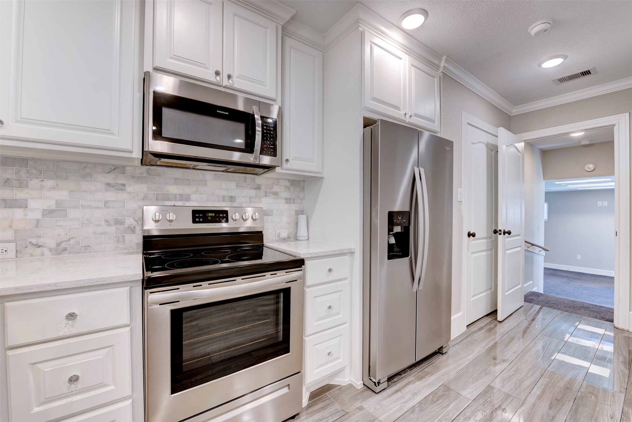 109 Dresden Street Houston, TX 77012 - Photo 18 of 43 Modern kitchen with stainless steel appliances, including a microwave and refrigerator. Features sleek white cabinetry, a marbled tile backsplash, and glossy wood-look flooring. Bright and welcoming with recessed lighting.