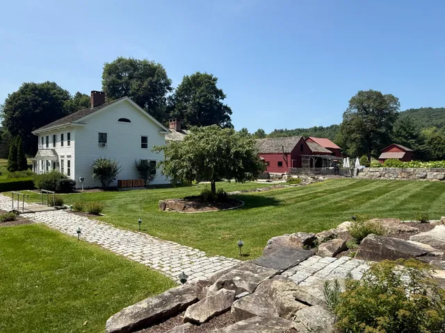 a view of a white house in front of a big yard with plants and large trees