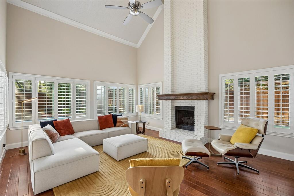 4115 Rive Lane Addison, TX 75001 - Photo 9 of 25 Vaulted ceiling living room featuring a floor-to-ceiling whitewashed brick fireplace with a wood mantle, wood-finish flooring, and multiple windows with plantation shutters