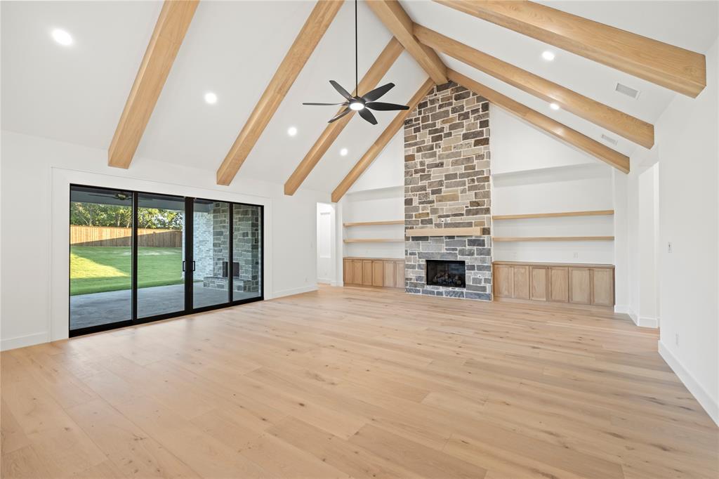 108 Bel Grand Road Haslet, TX 76052 - Photo 15 of 36 Vaulted ceiling with exposed wood beams, stone fireplace with a built-in hearth, wood-finish flooring, built-in shelving, and sliding glass doors opening to a concrete patio and manicured lawn