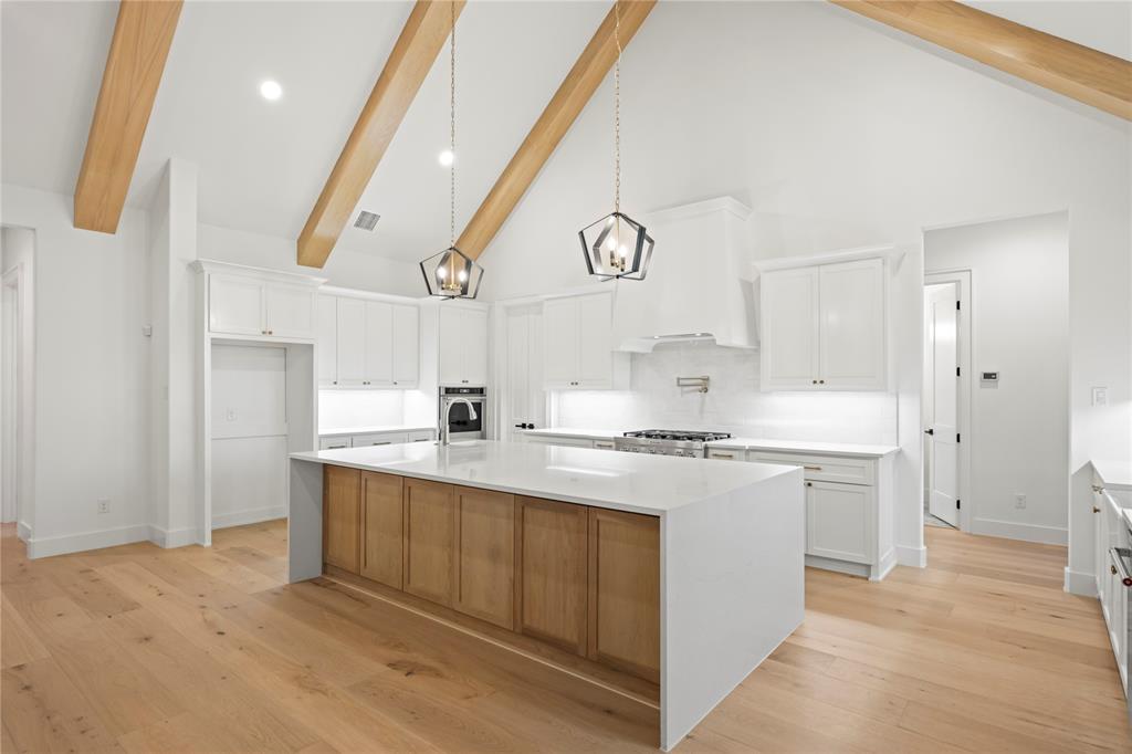 108 Bel Grand Road Haslet, TX 76052 - Photo 20 of 36 Vaulted ceiling kitchen featuring exposed wood beams, a spacious central island with a waterfall countertop, white shaker cabinetry, light wood-finish flooring, and two modern pendant lights