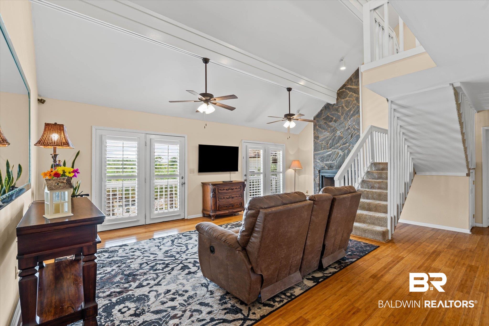 Living room featuring beam ceiling, light wood-type flooring, a stone fireplace, stairway, and high vaulted ceiling