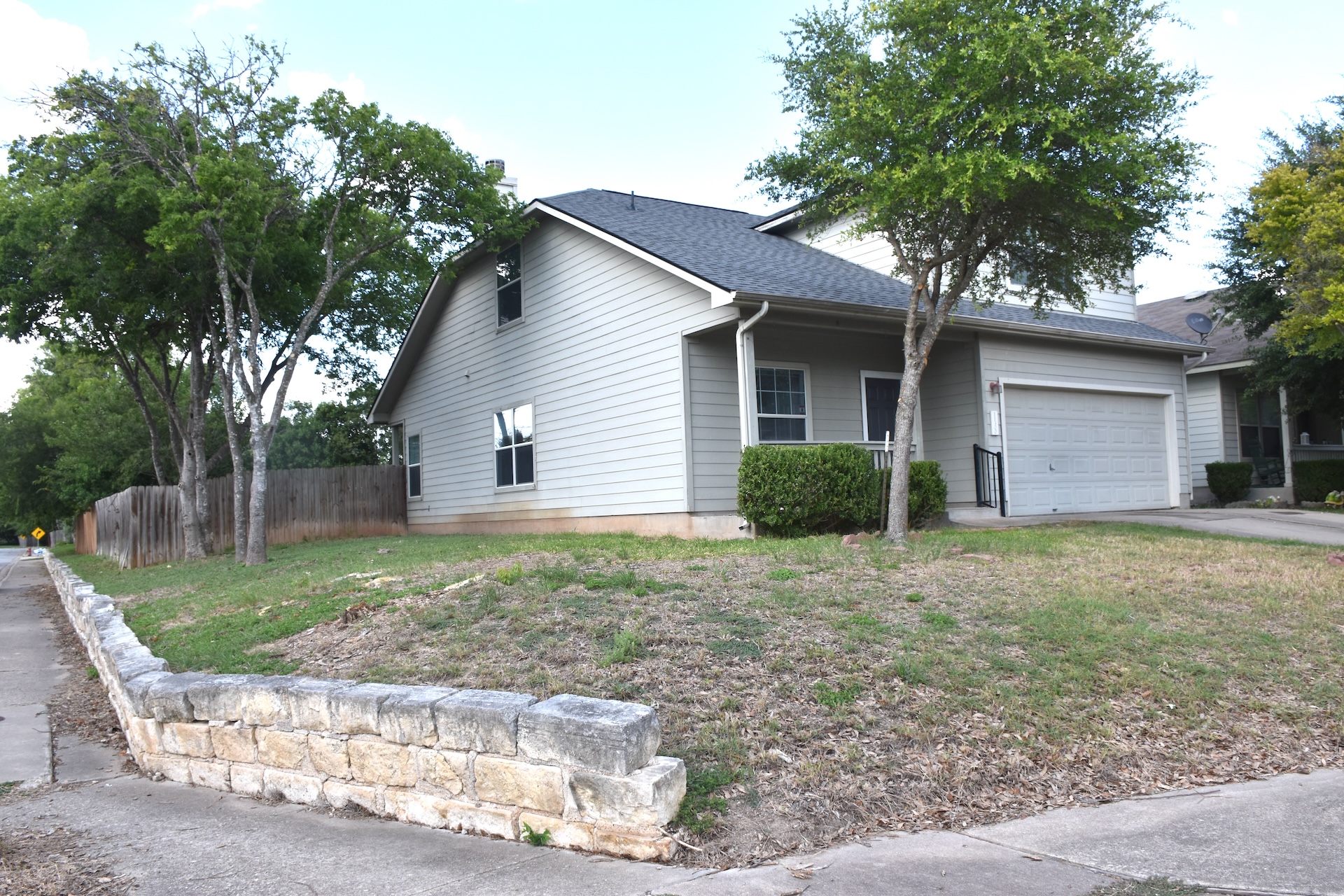 11300 Dimmit Street Manor, TX 78653 - Photo 2 of 23 A classic gray-sided residence with a single-car garage, complemented by mature trees and a charming stone wall.