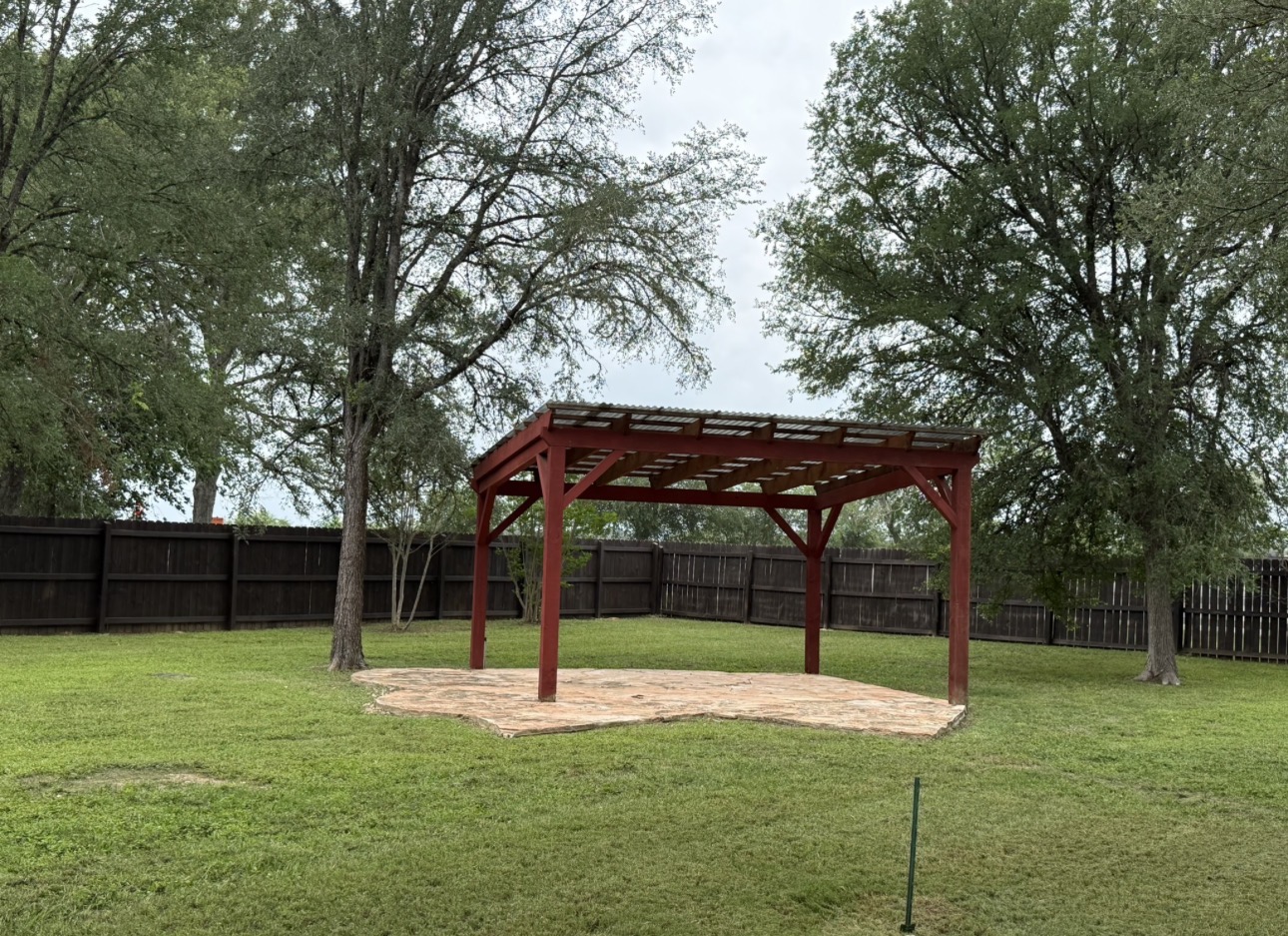 104 Spring Branch Bastrop, TX 78602 - Photo 7 of 32 A spacious backyard with mature trees and a charming red pergola over a patterned patio.
