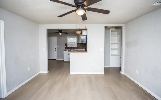 an empty room with wooden floor a ceiling fan and kitchen view